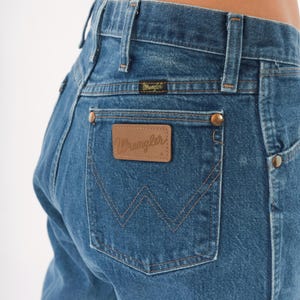 Puede incluir: Primer plano de vaqueros azules con el logo de Wrangler en el bolsillo trasero. Los vaqueros tienen un dise&ntilde;o cl&aacute;sico de cinco bolsillos con remaches de color cobre y un parche de cuero. Las costuras son de color beige.