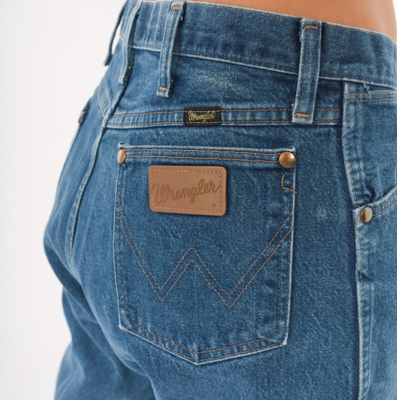 Puede incluir: Primer plano de vaqueros azules con el logo de Wrangler en el bolsillo trasero. Los vaqueros tienen un dise&ntilde;o cl&aacute;sico de cinco bolsillos con remaches de color cobre y un parche de cuero. Las costuras son de color beige.