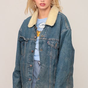 Puede incluir: Una chaqueta de mezclilla azul con cuello de sherpa color crema, mangas largas y cierre frontal con botones de metal. La chaqueta incluye dos bolsillos en el pecho con solapas abotonadas y se lleva sobre una camiseta azul claro con estampado.