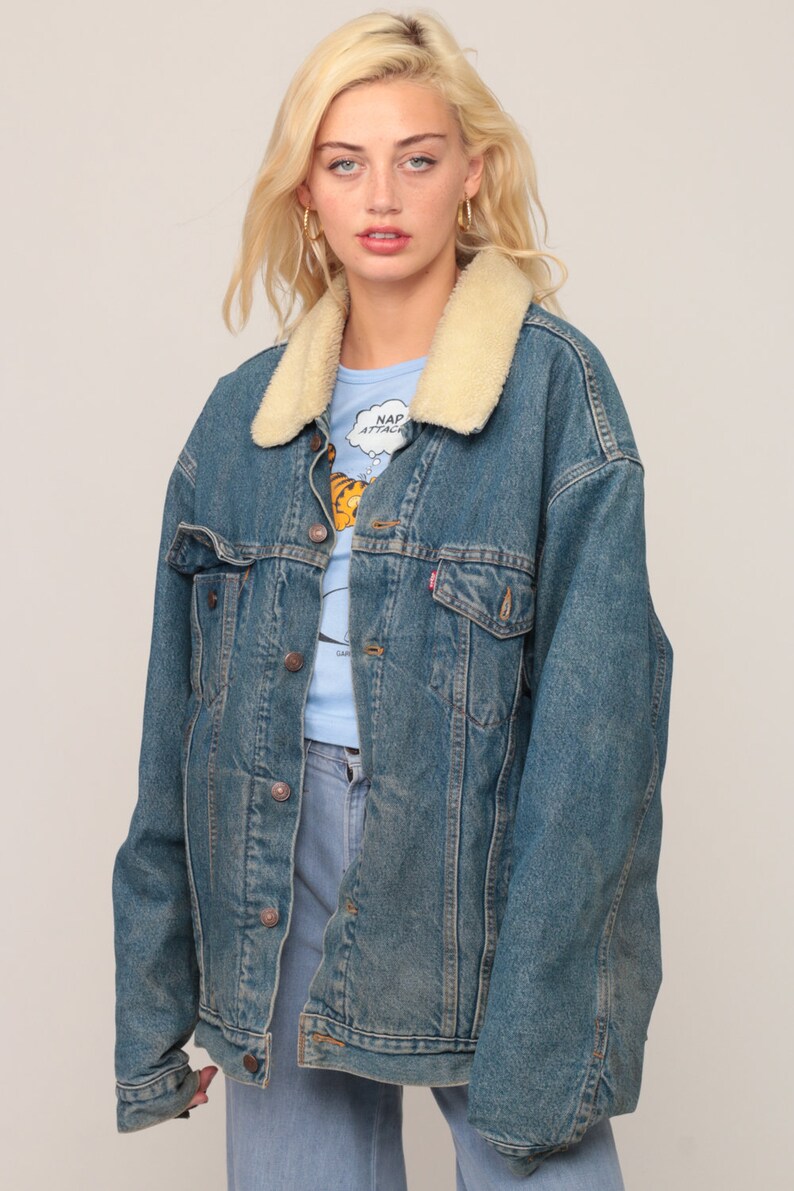 Puede incluir: Una chaqueta de mezclilla azul con cuello de sherpa color crema, mangas largas y cierre frontal con botones de metal. La chaqueta incluye dos bolsillos en el pecho con solapas abotonadas y se lleva sobre una camiseta azul claro con estampado.