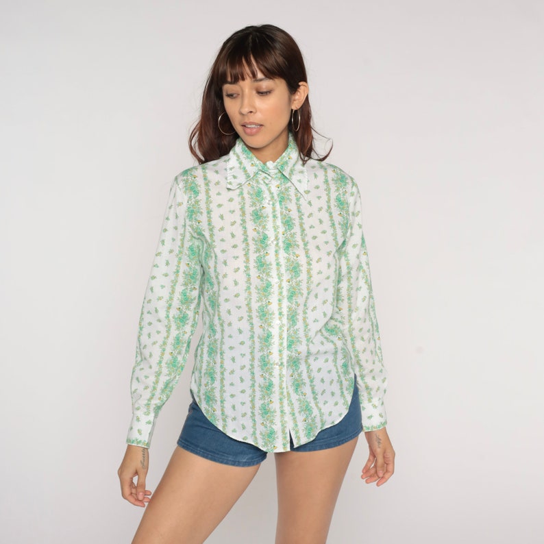 Puede incluir: Una camisa blanca de manga larga con botones y un estampado floral verde y blanco. La camisa tiene cuello y cierre de botones en la parte delantera.
