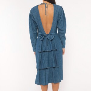 Puede incluir: Un vestido de mezclilla azul de tono medio con mangas largas y un dise&ntilde;o de espalda abierta profunda. La parte posterior incluye un cierre de lazo en el cuello y un gran lazo de tela en la cintura, sobre tres capas de volantes de mezclilla.