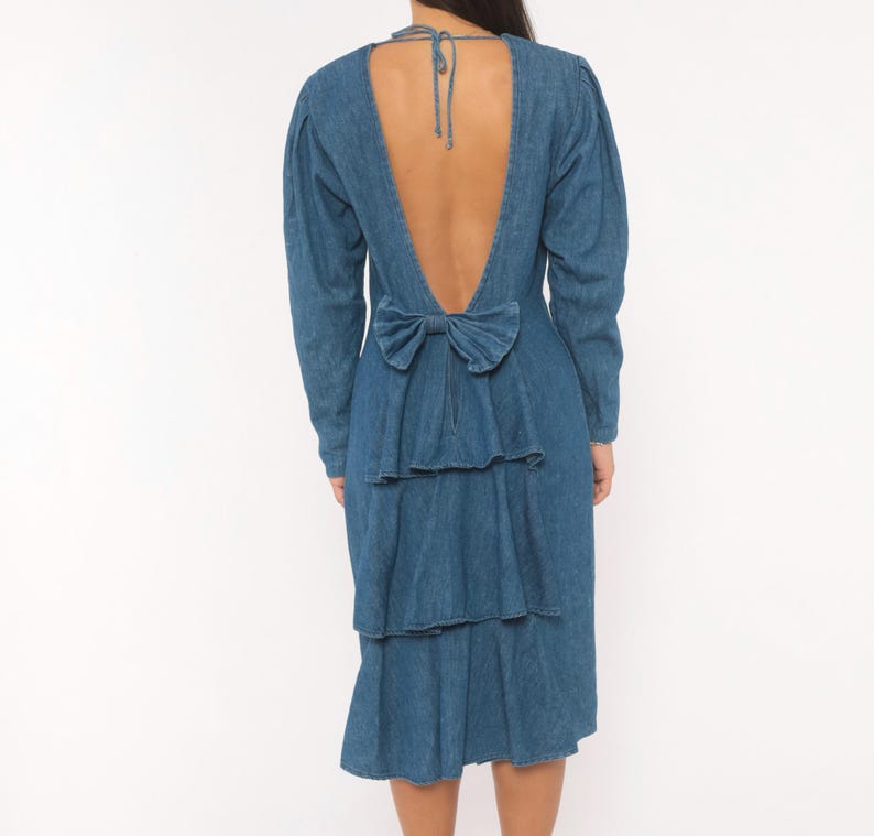Puede incluir: Un vestido de mezclilla azul de tono medio con mangas largas y un dise&ntilde;o de espalda abierta profunda. La parte posterior incluye un cierre de lazo en el cuello y un gran lazo de tela en la cintura, sobre tres capas de volantes de mezclilla.