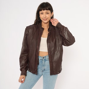 May include: A dark brown leather jacket with a zip-up front, a stand-up collar, and long sleeves. The jacket has a slightly textured appearance and a classic bomber jacket style. The model is wearing light blue jeans and a white top.