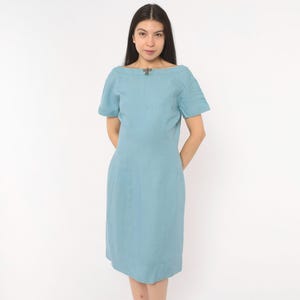 May include: A light blue knee-length dress with short sleeves and a boat neckline. The garment features a small, dark metallic decorative accent centered at the collarbone area. The dress has a straight, tailored silhouette with a simple hemline.
