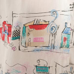 May include: A white shirt featuring a colorful hand-drawn style print of buildings and street scenes. The design includes black ink outlines with accents of red, blue, green, and pink. Text visible includes "CHEQUERS" and "THREE HORSE SHOES".