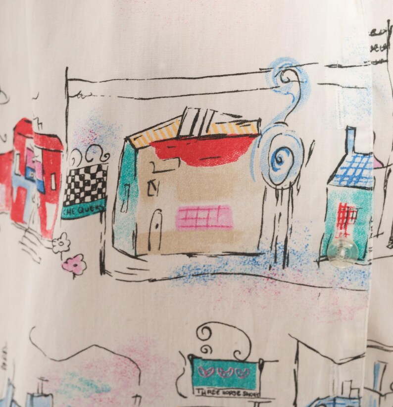 May include: A white shirt featuring a colorful hand-drawn style print of buildings and street scenes. The design includes black ink outlines with accents of red, blue, green, and pink. Text visible includes "CHEQUERS" and "THREE HORSE SHOES".