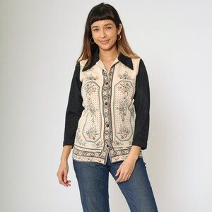 May include: Cream-colored button-down shirt with a floral pattern and black sleeves. The shirt has a pointed collar and is paired with blue jeans. The floral pattern includes orange, gray, and green accents.
