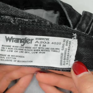 May include: Close-up of black Wrangler jeans with a white label. The label shows the brand name, size 76 x 91 cm, and washing instructions. The jeans are made of 100% cotton.
