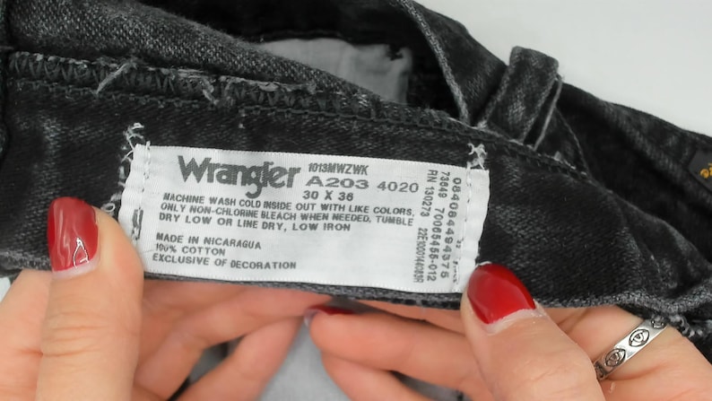 May include: Close-up of black Wrangler jeans with a white label. The label shows the brand name, size 76 x 91 cm, and washing instructions. The jeans are made of 100% cotton.