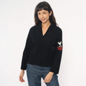 May include: Navy blue wool sweater with a V-neck collar. The long-sleeved sweater features an embroidered emblem of a white eagle and red stripes on the left sleeve. The sweater is paired with blue jeans.