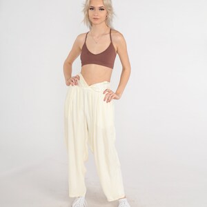May include: A woman wearing a brown ribbed bralette and cream-colored, loose-fitting pants. The pants have a twisted waist detail. White sneakers complete the outfit. The background is plain white.