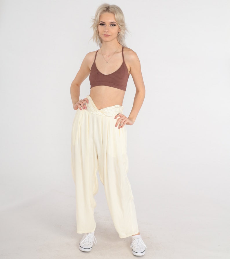 May include: A woman wearing a brown ribbed bralette and cream-colored, loose-fitting pants. The pants have a twisted waist detail. White sneakers complete the outfit. The background is plain white.