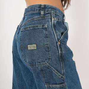 May include: Close-up of blue denim jeans with a square back pocket and a small rectangular label. The jeans have a classic five-pocket design with a carpenter-style pocket on the side. The denim is a medium wash.