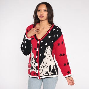 May include: A red, black, and white cardigan sweater featuring a Dalmatian dog design. The sweater has a V-neck, long sleeves, and a button-up front. The design includes paw prints and white polka dots.