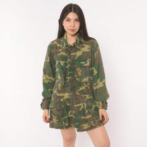 May include: A long-sleeve button-up shirt featuring a green, brown, and black camouflage pattern. The shirt includes two chest pockets with flaps and two larger pockets with flaps at the bottom hem, along with buttoned cuffs on the sleeves.