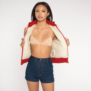 May include: A red and white zip-up vest with a faux fur lining. The vest is unzipped and the person is wearing a nude bra and dark blue denim shorts.