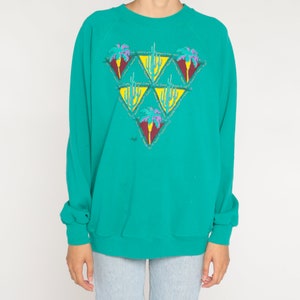 May include: Teal-colored sweatshirt with a desert-themed graphic. The graphic features a series of yellow triangles, each containing a cactus or palm tree design in shades of red, purple, and green. The sweatshirt has long sleeves and a crew neck.