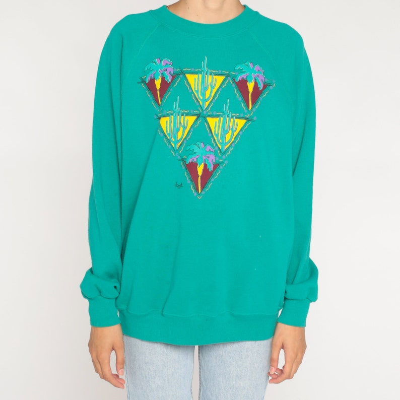 May include: Teal-colored sweatshirt with a desert-themed graphic. The graphic features a series of yellow triangles, each containing a cactus or palm tree design in shades of red, purple, and green. The sweatshirt has long sleeves and a crew neck.