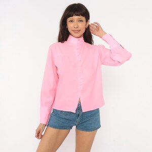 May include: A bright pink long-sleeved button-down shirt featuring a high mock neck collar, a front placket with matching fabric-covered buttons, and wide cuffs. The shirt is paired with blue denim shorts that have a classic five-pocket design.