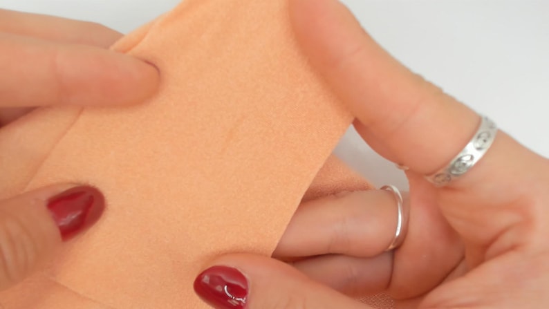 Puede incluir: Primer plano de tela color melocot&oacute;n sostenida entre los dedos, con esmalte de u&ntilde;as rojo y anillos plateados. La tela parece suave, posiblemente parte de una prenda o proyecto de manualidades.