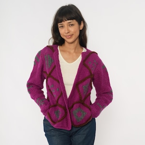May include: A magenta knit cardigan with a geometric pattern in gray and brown. The open-front sweater has a hood and pockets. The model is wearing a white shirt and blue jeans.