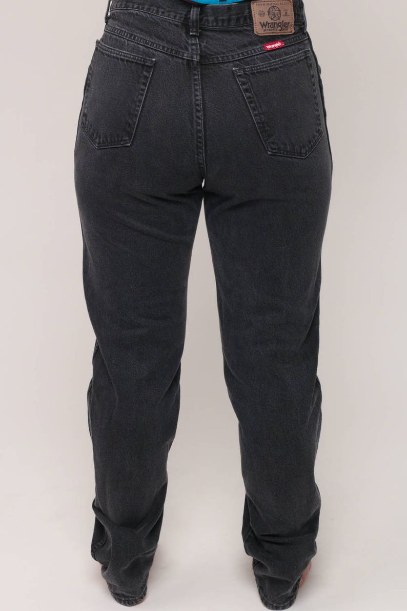 May include: Dark gray Wrangler jeans with a classic five-pocket design. The jeans have a straight leg fit and a Wrangler brand patch on the back pocket. The denim appears to be pre-worn.