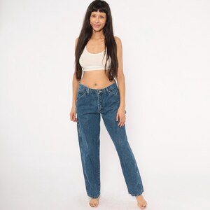 May include: Blue denim carpenter jeans with a straight leg fit. The jeans have a classic five-pocket design and a button and zipper closure. The model is wearing a white crop top.