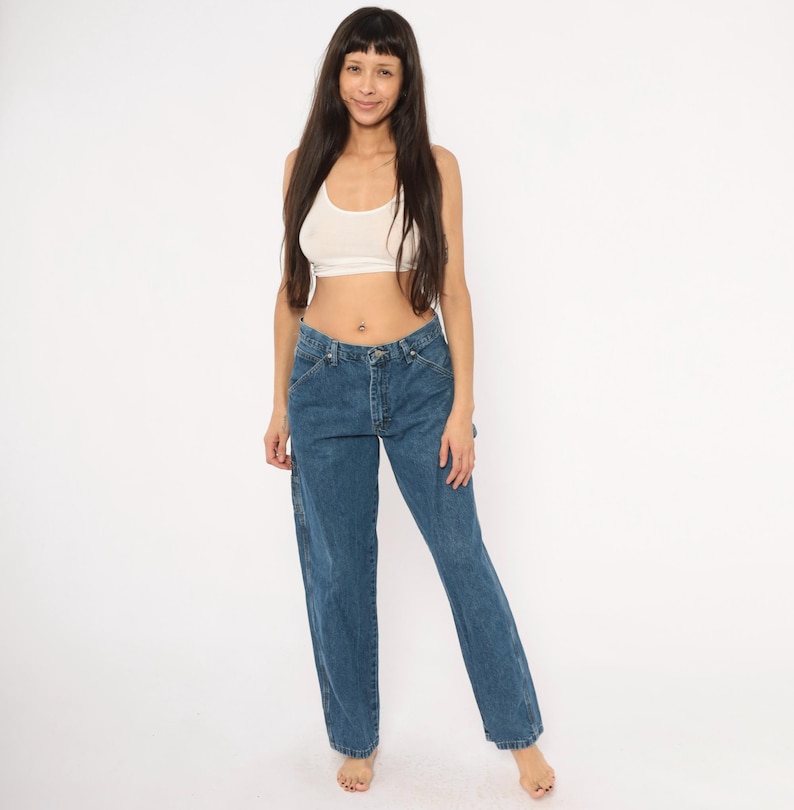 May include: Blue denim carpenter jeans with a straight leg fit. The jeans have a classic five-pocket design and a button and zipper closure. The model is wearing a white crop top.