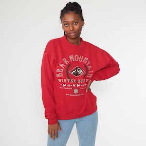 May include: A red sweatshirt with the words "Bear Mountain Winter Sprt" in white and green lettering. The sweatshirt has a crew neck and long sleeves. The graphic includes a bear, a diamond shape, and snowflakes.