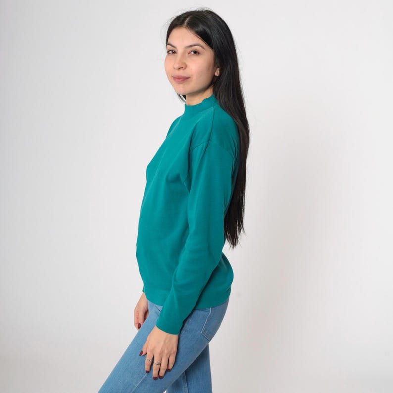 Pode incluir: Camisola verde-azulado de manga comprida com gola alta. A camisola &eacute; combinada com jeans azul claro. A modelo est&aacute; virada para a c&acirc;mara, com cabelo comprido e preto.