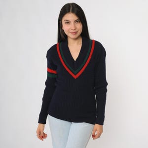 May include: Navy blue V-neck sweater with red and green stripes on the collar and sleeve. The sweater has a ribbed texture and long sleeves. A classic, sporty style.
