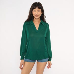 May include: A long-sleeved emerald green shirt featuring a pointed collar, a V-neckline, and cuffed sleeves. The shirt is paired with blue denim shorts. The fabric has a smooth, slightly reflective finish, and the design includes a simple, clean cut.