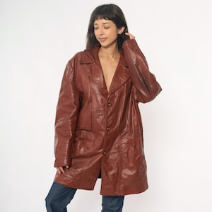 May include: A vintage brown leather coat with a notched collar, button closure, and front pockets. The coat has a longline silhouette and a slightly oversized fit. The leather has a worn, textured appearance. A classic outerwear piece.