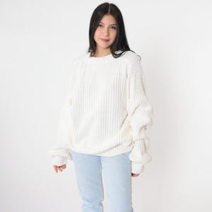 May include: A cream-colored, ribbed knit sweater with long sleeves. The sweater has a relaxed fit and a crew neckline. The model is wearing light blue jeans. The background is plain white.