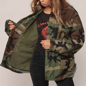 May include: A camouflage field jacket featuring a woodland pattern in shades of green, brown, and black. The jacket has a front zipper closure, a high collar, and a circular patch on the left chest. The interior lining is a solid olive green color.