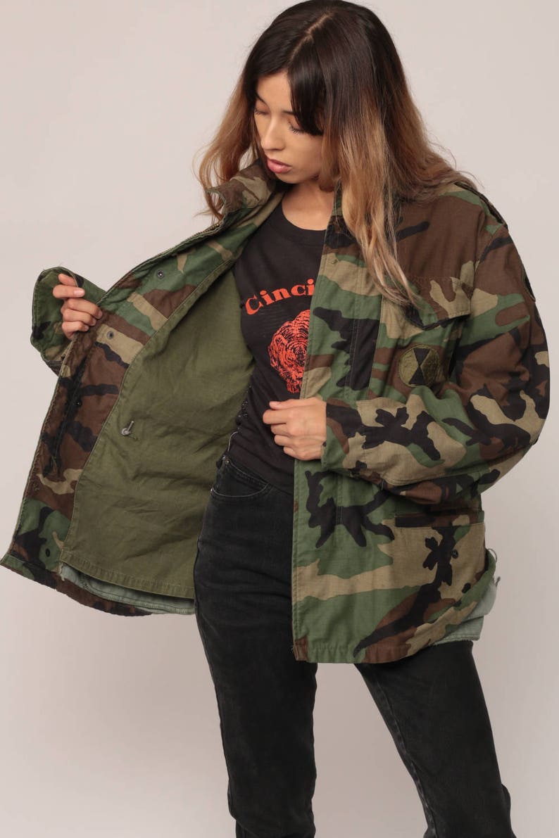 May include: A camouflage field jacket featuring a woodland pattern in shades of green, brown, and black. The jacket has a front zipper closure, a high collar, and a circular patch on the left chest. The interior lining is a solid olive green color.