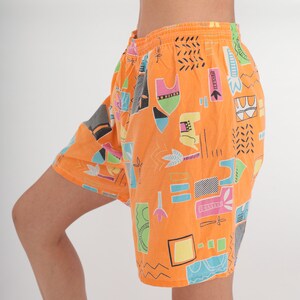 Op de afbeelding: Oranje shorts met een kleurrijk geometrisch patroon. Het patroon bevat abstracte vormen, lijnen en swirls in tinten roze, blauw, groen en zwart.