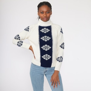 May include: White turtleneck sweater with a blue and white geometric pattern. The sweater has a ribbed texture and a relaxed fit.