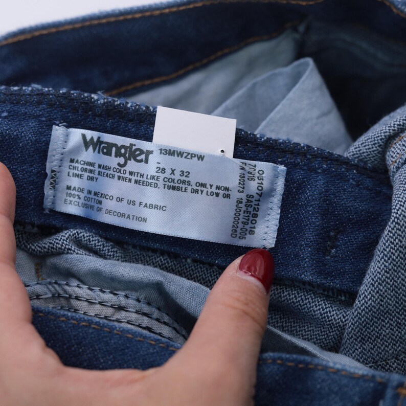 May include: Close-up of a Wrangler jeans label. The label is white with blue text and includes the Wrangler logo, size 28 x 32, and washing instructions. The jeans are a dark blue denim color.