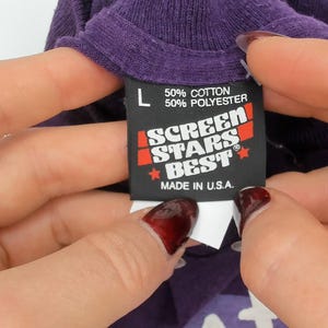 May include: Close-up of a purple t-shirt collar with a black tag. The tag reads "L", "50% Cotton 50% Polyester", and "SCREEN STARS BEST MADE IN U.S.A." The tag is held by two hands with rings.