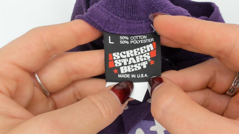 May include: Close-up of a purple t-shirt collar with a black tag. The tag reads "L", "50% Cotton 50% Polyester", and "SCREEN STARS BEST MADE IN U.S.A." The tag is held by two hands with rings.