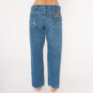Pode incluir: Jeans de ganga azul claro com um corte reto cl&aacute;ssico. Os jeans apresentam um remendo de couro castanho no bolso traseiro e uma lavagem desbotada. A vista traseira mostra os bolsos e as presilhas.