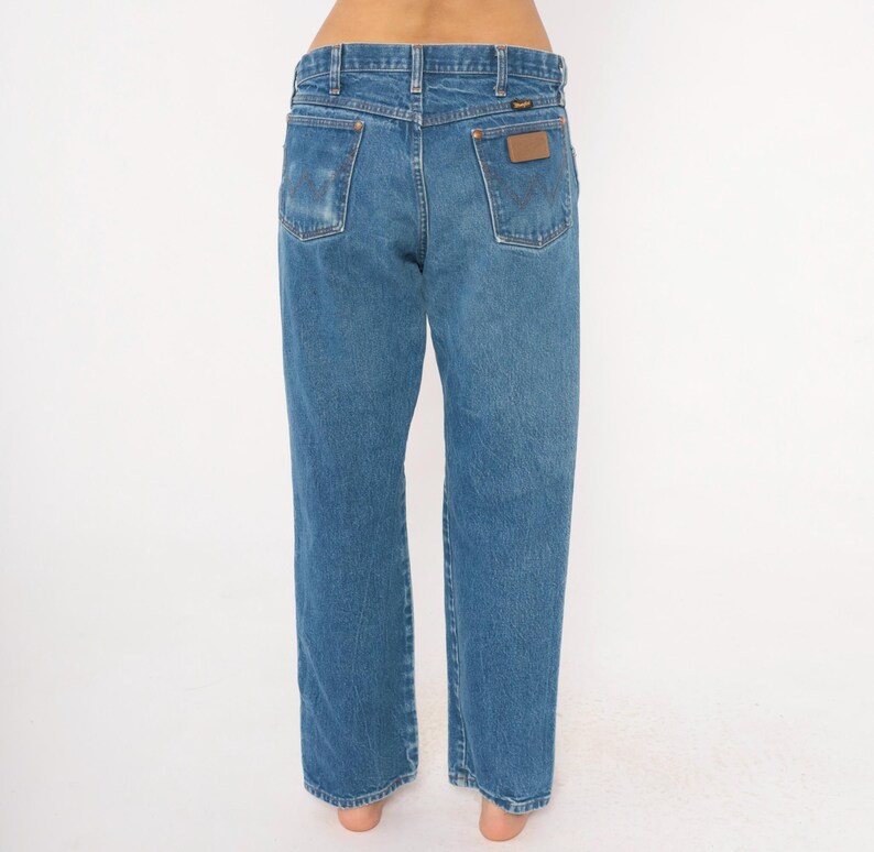 Pode incluir: Jeans de ganga azul claro com um corte reto cl&aacute;ssico. Os jeans apresentam um remendo de couro castanho no bolso traseiro e uma lavagem desbotada. A vista traseira mostra os bolsos e as presilhas.