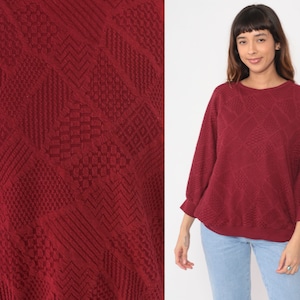 Puede incluir: Un suéter color granate con un patrón geométrico texturizado. El suéter tiene cuello redondo y mangas largas.