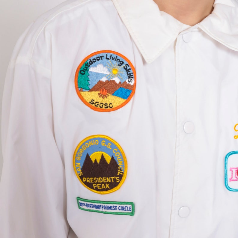 Op de afbeelding: Wit overhemd met knopen en geborduurde patches. De patches tonen een berglandschap met de tekst "Outdoor Living Skills", een berglandschap met de tekst "San Gorgonio G.S. Council President's Peak" en de tekst "80th Birthday Promise Circle".