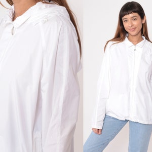 May include: White hooded jacket with a full-length zipper closure. The jacket has long sleeves and a collared neckline. The fabric appears lightweight and suitable for layering. The jacket is shown on a person wearing blue jeans.
