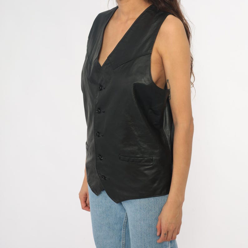 May include: Black leather vest with a V-neck, button closure, and two front pockets. The vest is sleeveless and appears to be vintage. The vest is worn over a light blue pair of jeans.