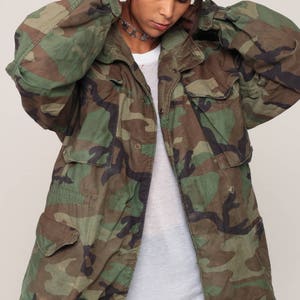 May include: A camouflage field jacket featuring a woodland pattern in shades of green, brown, and black. The jacket includes a front button closure, a fold-over collar, and four large flap pockets on the front, worn over a plain white t-shirt.