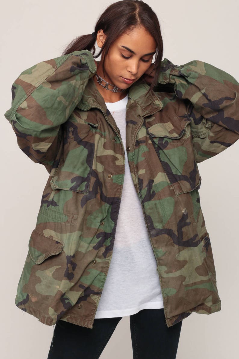 May include: A camouflage field jacket featuring a woodland pattern in shades of green, brown, and black. The jacket includes a front button closure, a fold-over collar, and four large flap pockets on the front, worn over a plain white t-shirt.